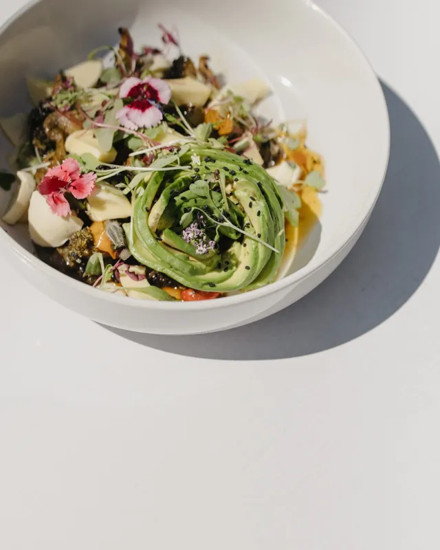 A bowl of avocado, greens and edible flowers on a white table, seen from above.